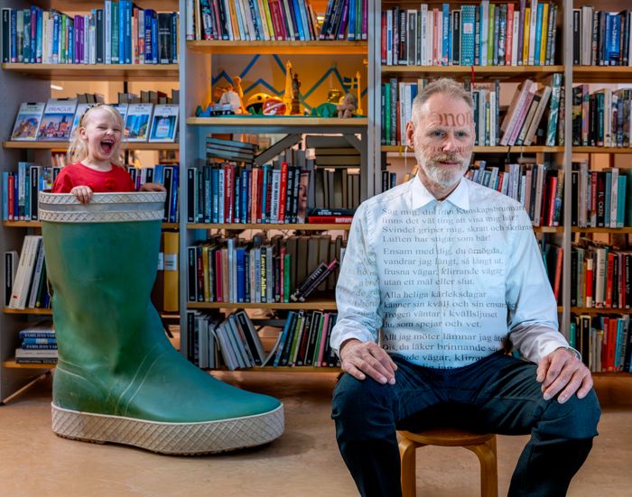 En man sitter i ett bibliotek med bokhyllor bakom sig. Till vänster om honom, ett barn som tittar upp ur en jättestor gummistövel.