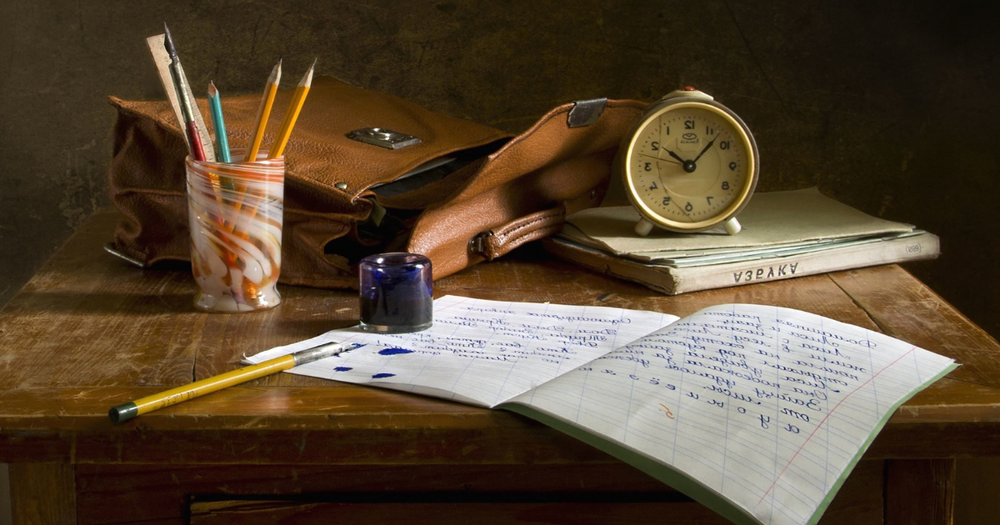 Bild på bord med skrivbok, pennor, klocka och väska.