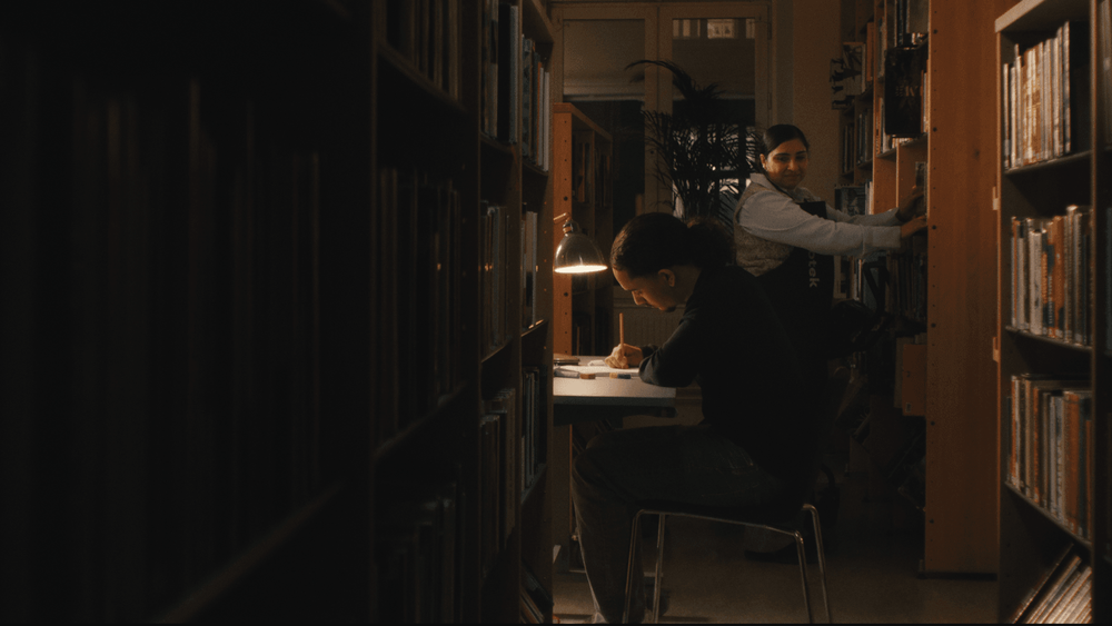 Interiör från bibliotek med kille som sitter och studerar och kvinna som sätter upp böcker.