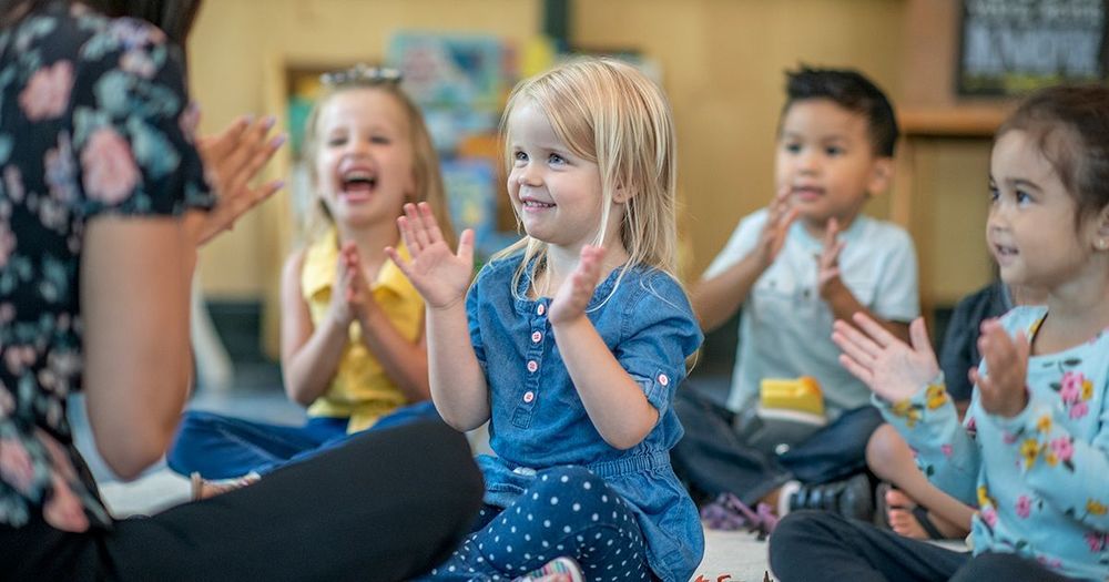 Barn i förskoleålder sitter på golvet och sjunger och klappar händerna tillsammans med en vuxen.
