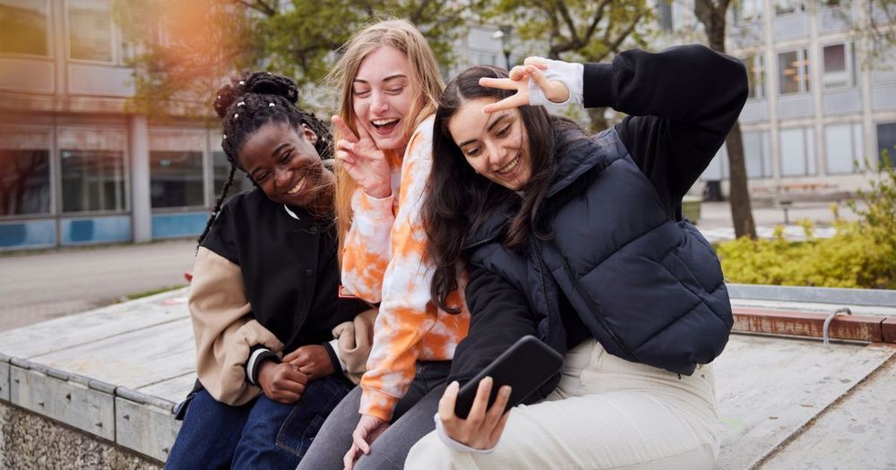 Tre unga tjejer sitter på ett torg och skrattar tillsammans.
